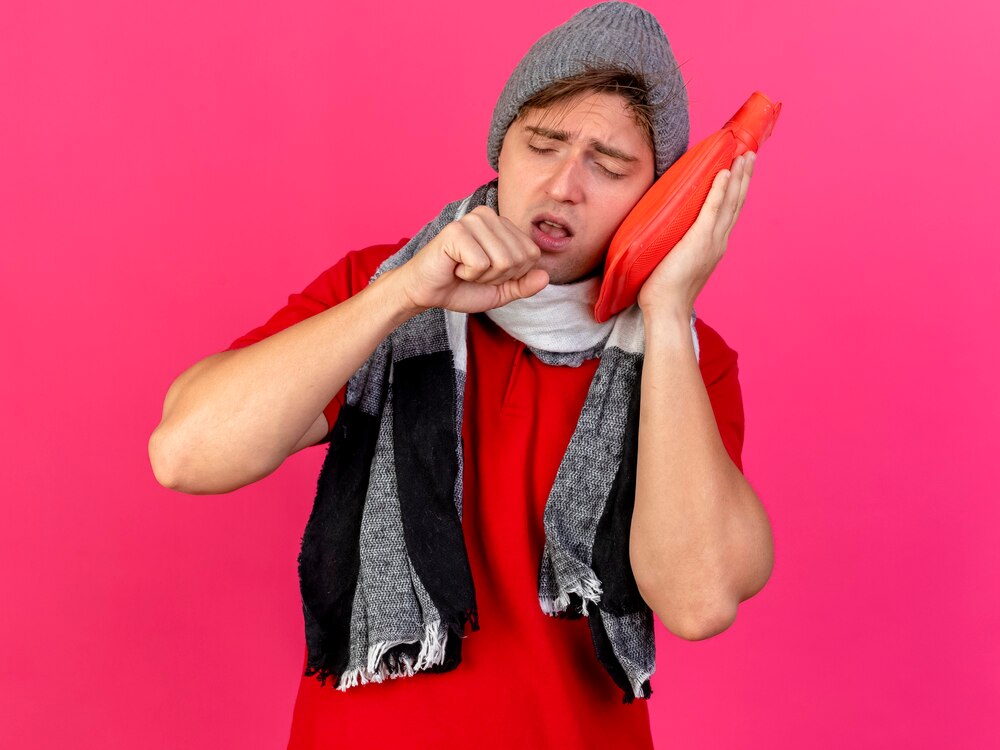 Young handsome blonde ill man wearing winter hat and scarf touching face with hot water bottle and coughing with closed eyes isolated on crimson background with copy space