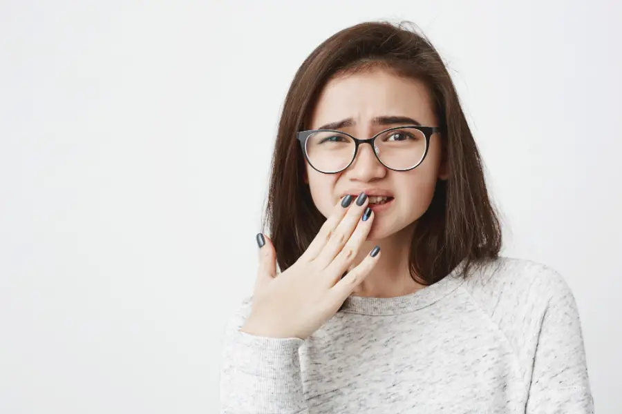Portrait of young cute female in glasses, squinting and covering her mouth with hand, feeling pain or sorry